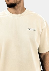Krémový sametový tričko s vroubkovaným kulatým výstřihem. Na levé straně hrudníku se nachází malé černé logo "CRYSTAL".
