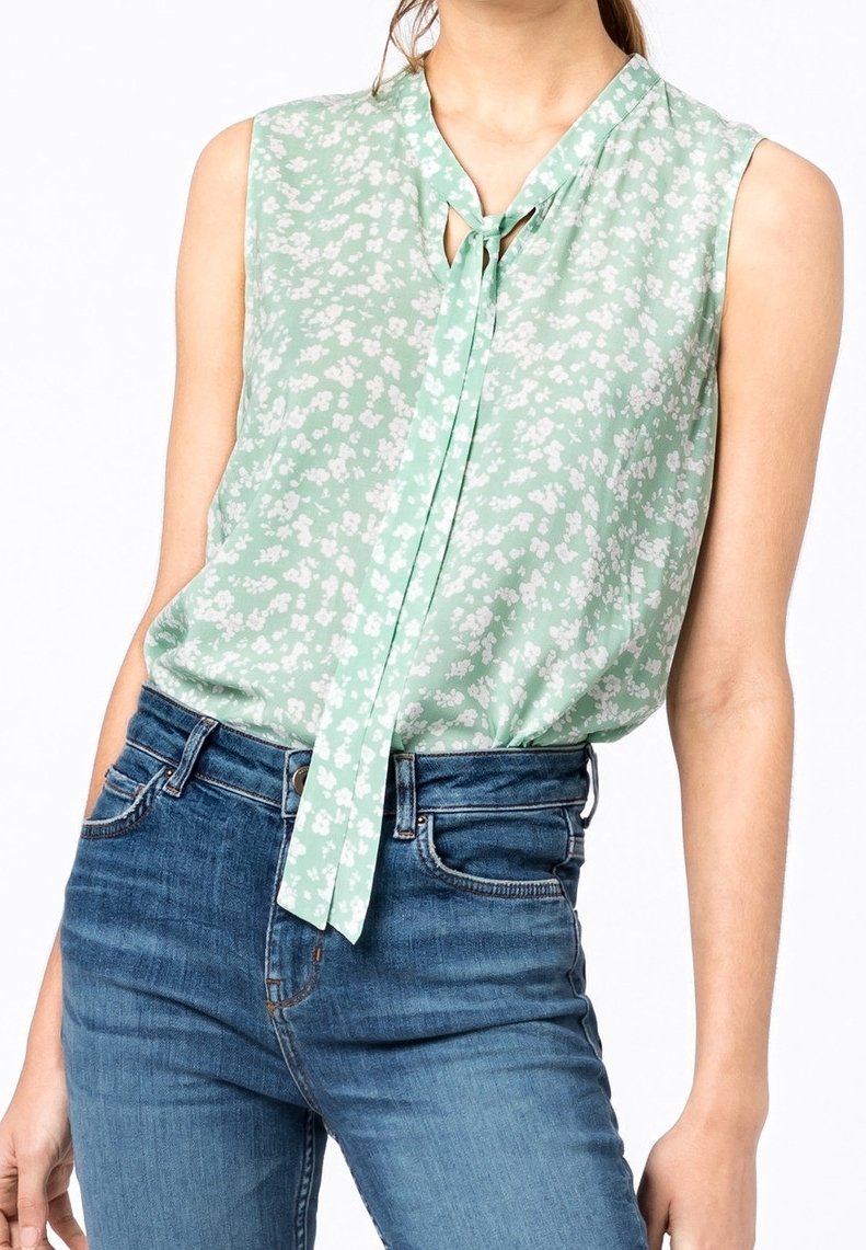 Blouse sans manches vert menthe à motif floral avec détail de nœud devant, portée rentrée dans un jean bleu taille haute par une personne.