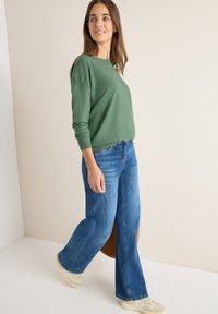 Maglione verde a maglia, vestibilità rilassata, abbinato a jeans svasati azzurri chiari. Gli accessori includono sneakers beige e una borsa marrone. Tessuto liscio.