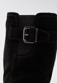 Bottine en daim noir avec une surface texturée, un panneau élastique et un accent de boucle métallique sur le côté. Doublure intérieure lisse.