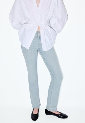 Persoon draagt lichtblauwe spijkerbroek, oversized wit overhemd met volumineuze mouwen en zwarte ballerina's, staand tegen een witte achtergrond.