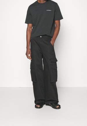 Czarna koszulka z krótkim rękawem z napisem "BRAK NARODOWOŚCI", w połączeniu z szerokimi czarnymi spodniami cargo z wieloma kieszeniami i luźnym krojem.