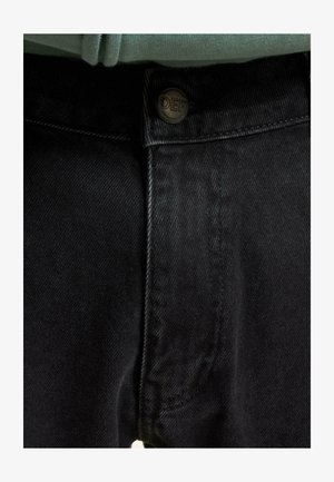 Musta denimpüksid eesnupuga, millel on tekst "DEF." Kangas on sileda tekstuuriga ja talje on veidi kitsenev.