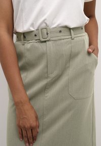 Vihreä hame, jossa on neliön muotoinen solki, sivutaskut ja teksturoitu kangas, yhdistettynä valkoiseen yläosaan.