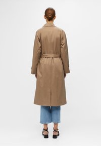 Mujer con pelo castaño recogido en un moño, lleva un abrigo trinchera color caqui a la altura de la rodilla, jeans azules cortos y sandalias negras de tacón, vista desde atrás.