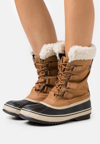 Jambes portant des bottes d'hiver isolées tan et noires avec doublure en polaire blanche et fermeture à lacets sur un fond blanc.
