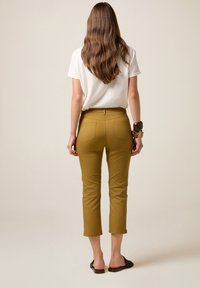 Mosterdgele, aansluitende broek met een gemiddelde taillehoogte, cropped lengte en twee achterzakken, gecombineerd met een wit T-shirt en zwarte sandalen.