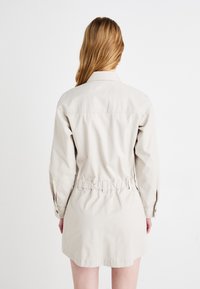 Beige bomull skjortekjole med lange ermer, en tilpasset midje, knappelukking og buet sømkant, som fremviser en jevn tekstur og et enkelt design.
