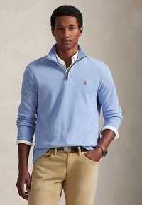 Hombre que lleva un jersey con cremallera hasta un cuarto en azul claro sobre camiseta blanca, pantalones color beige y reloj marrón en la muñeca, de pie con una mano en el bolsillo.