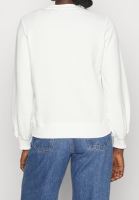 Felpa bianca con vestibilità rilassata, maniche lunghe a palloncino, polsini e orlo a coste. Indossata con jeans di denim blu a gamba rastremata, con schiena scoperta.