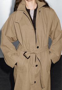 Beige trenchcoat van lichtgewicht stof, met een ceintuur in de taille, grote kraag en knoopdetails, en lange, relaxte mouwen.