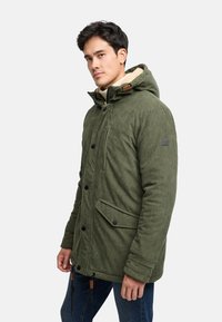 Olivgrüne Corduroy-Parka mit einer mit Kunstfell gefütterten Kapuze, schwarzen Knöpfen und Fronttaschen. Verfügt über Zugbänder am Saum zur Anpassung.