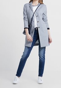 Veste grise à trois boutons, associée à un t-shirt blanc et un jean bleu foncé. Des baskets blanches complètent la tenue.