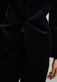 Giacca e pantaloni in velluto nero con vestibilità su misura, indossati da una persona, che mostra parte della mano e del tronco.