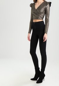 Metallisch goldene Wickelbluse mit langen Ärmeln und Schulterrüsche, kombiniert mit schwarzen Skinny Jeans und schwarzen Stiletto-Ankle-Boots.