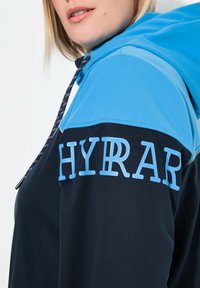 Huppari, jossa on sininen ja tummansininen suunnittelu. Hihan teksti lukee "HYRAR" vaaleansinisellä. Pehmeä rakenne ja muotoiltu istuvuus.