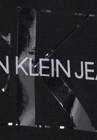 Schwarzes T-Shirt mit auffälligem weißen und silbernen Folien-Druck. Das Design enthält "CALVIN KLEIN JEANS" in Großbuchstaben.