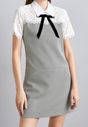 Frau trägt ein kurzärmeliges schwarz-weißes Hahnentrittmuster-Kleid mit weißer Spitze am Oberteil, weißem Kragen und schwarzer Samtschleife.