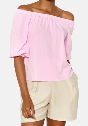 Donna che indossa una blusa rosa chiaro scollata con maniche fino al gomito e pantaloncini beige, in piedi con una mano in tasca.