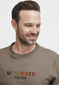 Sudadera marrón con tejido texturizado, cuello acanalado y bordado colorido: "MCMXXIV FQ1924" en verde, naranja y rojo.