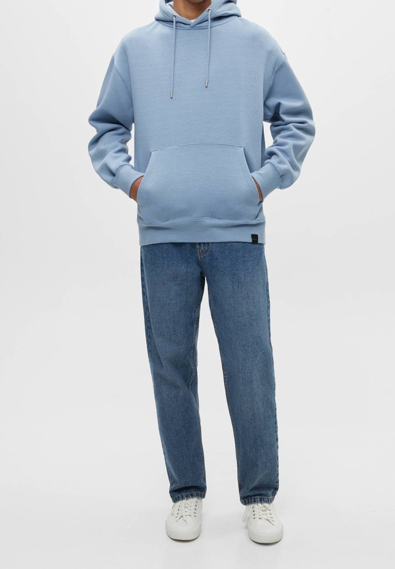 Personne portant un sweat à capuche bleu clair, un jean bleu et des baskets blanches, debout avec les mains dans les poches du sweat à capuche devant un fond uni.