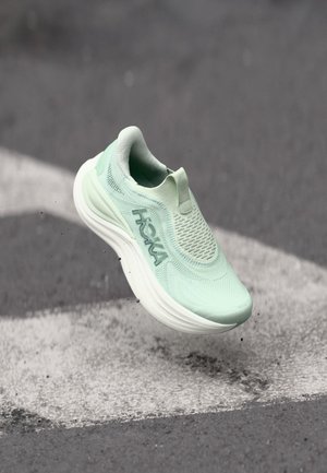Zapatilla de correr Hoka verde claro en el aire sobre asfalto con una señal de flecha blanca en el suelo.