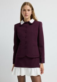 Veste bordeaux avec col blanc, sept boutons, deux poches avant et poignets texturés. Assortie à une mini-jupe blanche évasée.