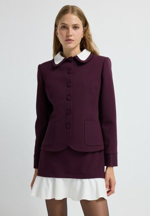 Maroon jacket with white collar, seven buttons, two front pockets, and textured cuffs. Paired with a flared white mini skirt.