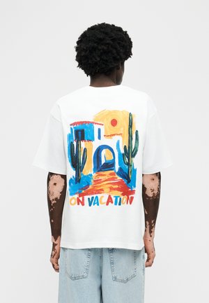 Oseba s temnimi kodrastimi lasmi in vitiligom nosi belo majico z barvitim prizorom puščave, kaktusi in napisom "NA POČITNICAH", ki je kombinirana s svetlimi kavbojkami.