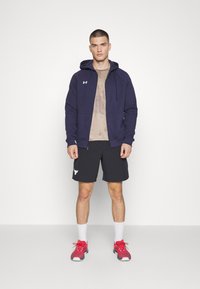 Marine blauwe zip-up hoodie over een lichtbruine t-shirt, gecombineerd met zwarte shorts met een logo. Rode en grijze sport schoenen met witte sokken.