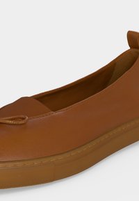 Καφέ δερμάτινο παπούτσι τύπου slip-on με στρογγυλεμένη μύτη, ελαστική ταινία στο πάνω μέρος, μικρή διακοσμητική φιόγκο και ελαστική σόλα σε λευκό φόντο.