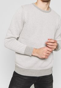 Mężczyzna w jasnoszarym, teksturowanym swetrze z prążkowanymi mankietami i dołem, poprawiający rękaw, w połączeniu z czarnymi dżinsami na jednolitym tle.