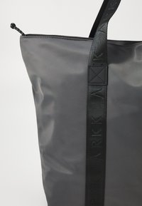 Čierna nylonová taška s hladkou textúrou, ktorá má potlačený popruh a zips. Dizajn je minimalistický a moderný.