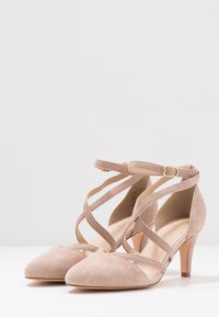 Zapatos de tacón alto de ante beige con punta afilada, tiras entrecruzadas y cierres de hebilla en el tobillo, sobre una superficie blanca y un fondo claro.