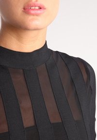 Mustapaita, jossa on korkea kaulus, läpikuultavaa kangasta ja pystysuoria mustia raitoja. Pintakuvio on sileä ja korostaa ihon alla.