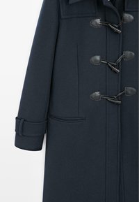 Cappotto di lana blu scuro con chiusura a toggle, colletto a revers e tasca laterale. Le maniche sono a polsino, mettendo in evidenza un motivo testurizzato.