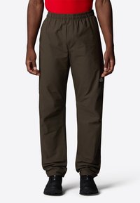 Pantalons légers marron avec une taille élastique, jambes droites et un détail de logo sur la cuisse gauche. Portés avec un haut rouge et des chaussures noires.