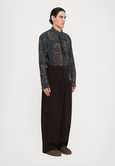 Camicia a maniche lunghe di colore verde scuro con un motivo floreale, abbinata a pantaloni larghi marroni e scarpe marroni. Nota: la camicia presenta un collo a cravatta.