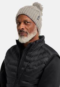 Berretto lavorato a maglia grigio con pom pom in cima, caratterizzato da una texture a costine e un piccolo etichetta del marchio. Indossato con un gilet isolante nero.