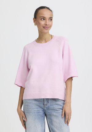 Mladá žena sa usmieva, má na sebe svetloružový krátky pletený sveter s krátkym rukávom a svetlomodré džínsy, stojí pred bielym pozadím.