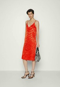 Modelo vestindo um vestido midi sem mangas com estampado de tigre vermelho, saltos pretos com tiras, e segurando uma bolsa com estampado de zebra em preto e branco.