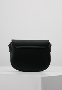 Černá kožená crossbody taška s kulatým tvarem, klopovým uzávěrem a bočními kovovými detaily. Hladký povrch, minimalistický design.