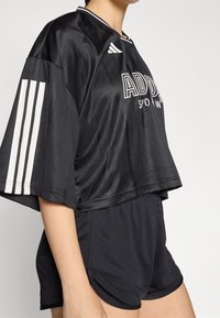 Černý sportovní cropped top s bílými pruhy na rukávech, obsahující logo "ADIDAS" a doplněný o černé šortky. Hladká, lehká tkanina.