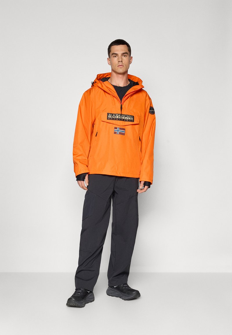 Giacca impermeabile arancione brillante con tasca frontale e logo, abbinata a pantaloni scuri. La giacca ha un cappuccio e polsini regolabili.