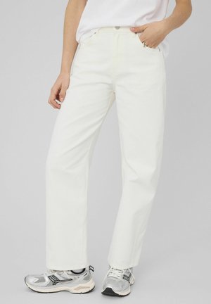 Personne portant un pantalon blanc taille haute à jambes droites, des chaussures de course argentées et une chemise blanche rentrée, main dans la poche.