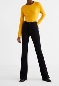 Maglione giallo a costine abbinato a jeans neri a vita alta e scarpe nere. L'outfit combina texture morbide e colori a contrasto.