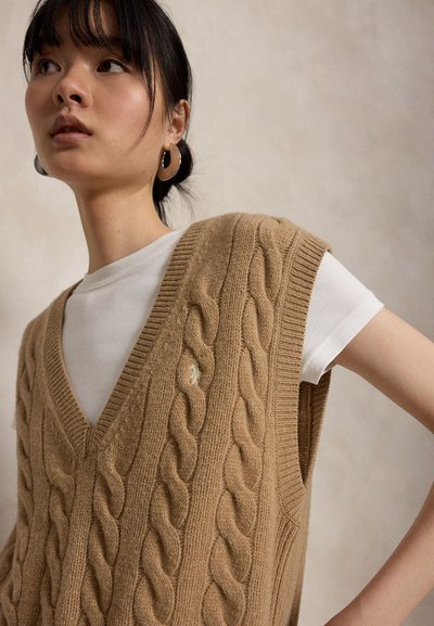 Ung kvinde med mørkt hår bærer en beige kabelstrikket ærmeløs sweater over en hvid skjorte, accessoriseret med store hoop-øreringe.