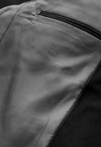 Közeli kép szürke texturált anyagról fekete varrással és keskeny vízszintes zsebélyukkal egy ruhadarabon.