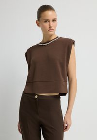 Hnědý zkrácený top s krátkými rukávy, se zaobleným výstřihem zdobeným korálkovým lemováním. Nosený s odpovídajícími přiléhavými hnědými kalhotami.