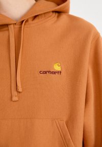 Orange hoodie made of soft fabric, featuring a front pocket and a drawstring hood. Embroidered Carhartt logo in yellow and burgundy.